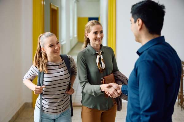 A teenage girl wearing a backpack smiles as her mother shakes hands with her teacher in the hallway of the school.