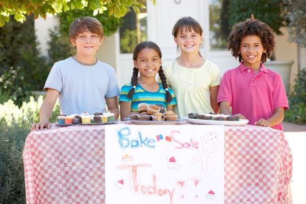 A group of children standing outside behind a bake sale table and smiling. There are muffins and cupcakes on the table.