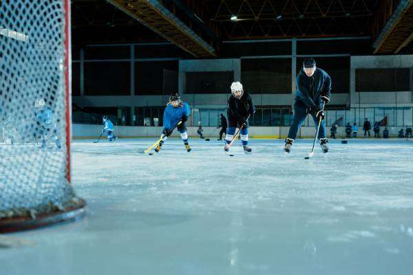 A hockey practice on an indoor rink. Players practice in the background while two young players skate toward the goal next to their coach.