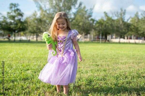 A little girl wearing a purple princess gown. She is standing in a field holding a stuffed animal of a chameleon.