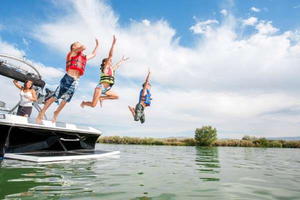 Two boys and a girl wearing life jackets and jumping off the back of a boat into a lake. Their mother is watching from the boat.