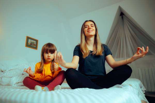 A young mother unwinds by practicing her daily yoga meditations with her daughter on the daughter's bed.