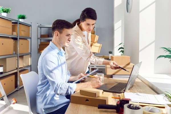 Two people working at a desk with a laptop on it. Several cardboard boxes neatly organized on shelves fill the background.