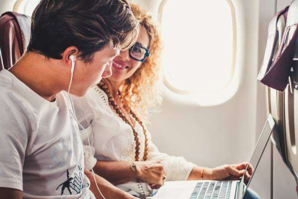 A woman sits by the window of an airplane with a laptop and looks over to a teen boy with headphones.