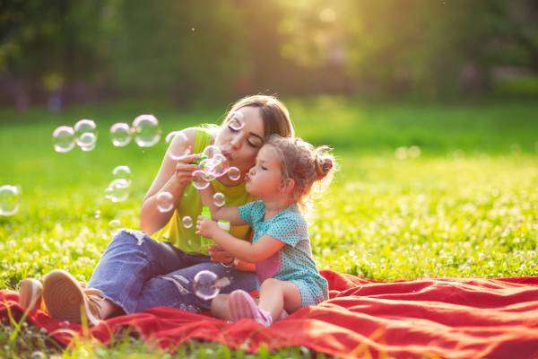 A young woman in a neon green shirt sitting on a red blanket with a little girl in polka dots, blowing bubbles together.