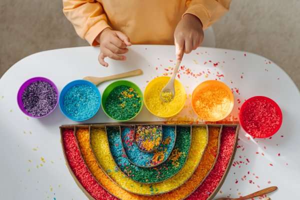 A child plays with red, orange, yellow, green, blue, and purple rice and places it in a rainbow-shaped container.