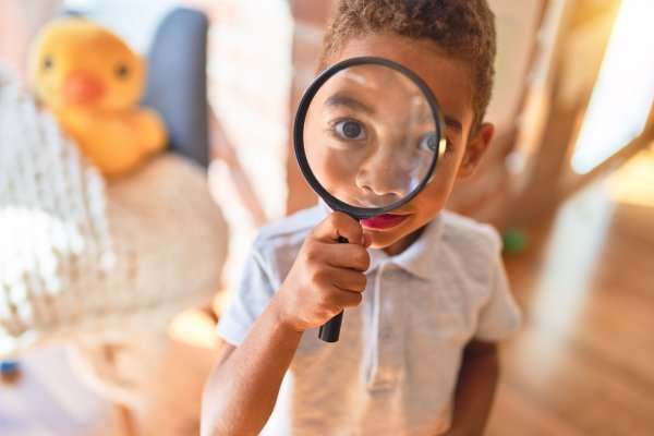 A child holds up and looks through a magnifying glass, which enlarges his eye. A duck toy sits behind him.