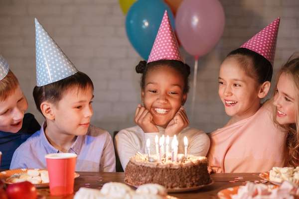 Small children in colorful, polka-dot party hats gathered around a birthday cake with multiple lit candles.