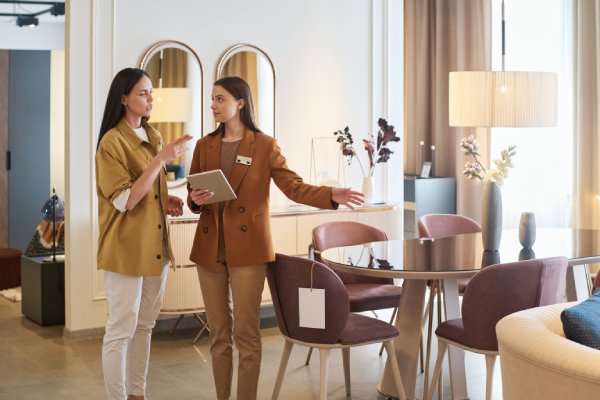 A female shopper standing with a female furniture store sales associate. The associate is pointing to a dining set.