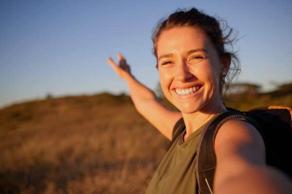 A young, smiling woman with a backpack taking a selfie outdoors and gesturing toward a grassy field.