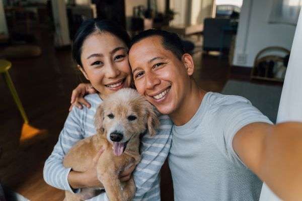 A smiling man and woman holding a happy dog in a warmly lit room with wooden floors and cozy furniture.