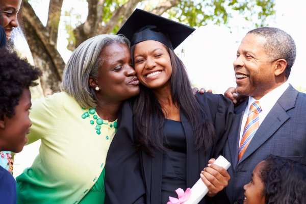 A young female in a black graduation gown and hat is being embraced by two parents in formal attire. Two children look on.