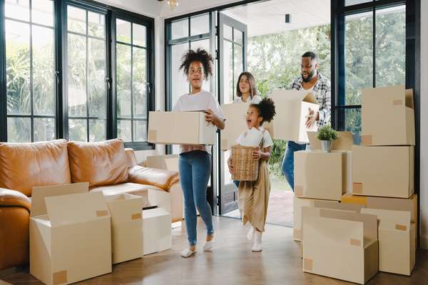 A family of two adults and two children is walking into a home with large windows and stacks of moving boxes.