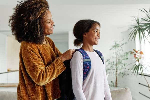 A mother and her daughter are smiling and talking as the mother helps her child put her backpack on for school.