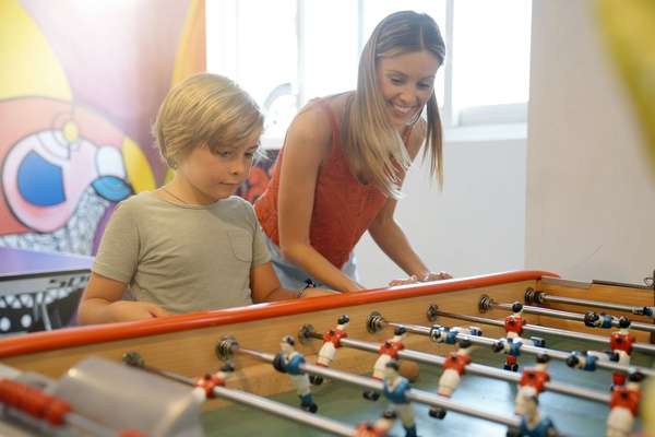 A woman and a young boy smile while playing foosball together at a table in a bright indoor recreation area.