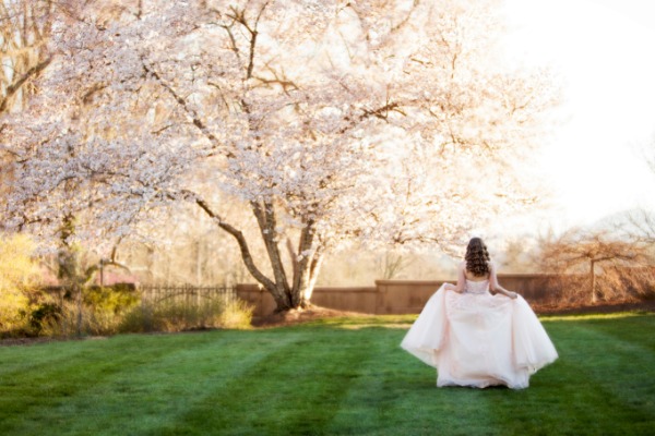 A teen girl in a pale formal dress walking across a grassy lawn toward blossoming trees in warm sunlight.