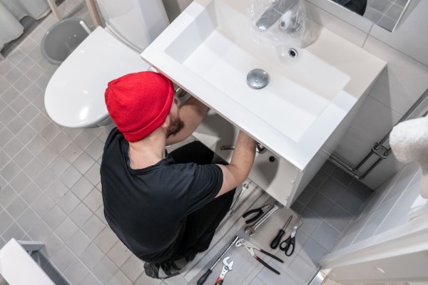A man in a beanie using tools to adjust something below a bathroom sink. The floor is littered with other tools.