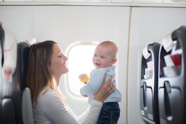 A young mother holding her baby in front of her as she sits on a plane and both smile at one another.