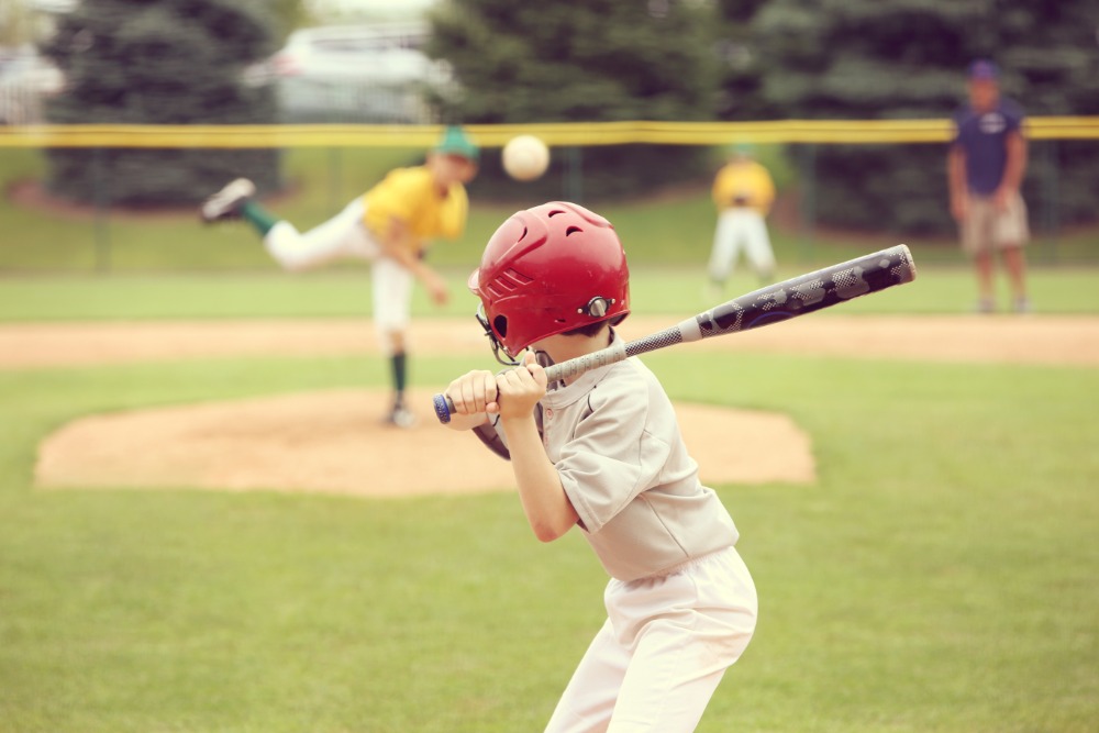 A view from behind of a young baseball player with a red helmet and black bat preparing for an incoming pitch.