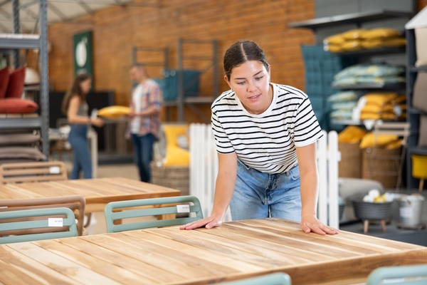 A woman in a striped shirt and jeans leans to touch a wooden outdoor table display in a furniture store.