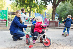 Happy female teacher assisting girl to ride tricycle on playground