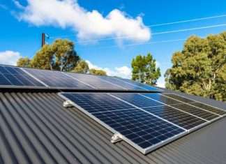 A metal roof with rows of solar panels on a partly-cloudy day. Trees and telephone wires are seen in the background.