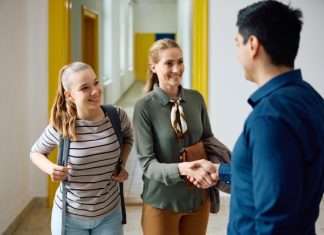 A teenage girl wearing a backpack smiles as her mother shakes hands with her teacher in the hallway of the school.