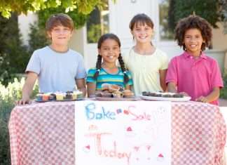 A group of children standing outside behind a bake sale table and smiling. There are muffins and cupcakes on the table.