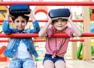 Two kids sitting on playground equipment with VR headsets. The equipment is red and the kids are smiling.