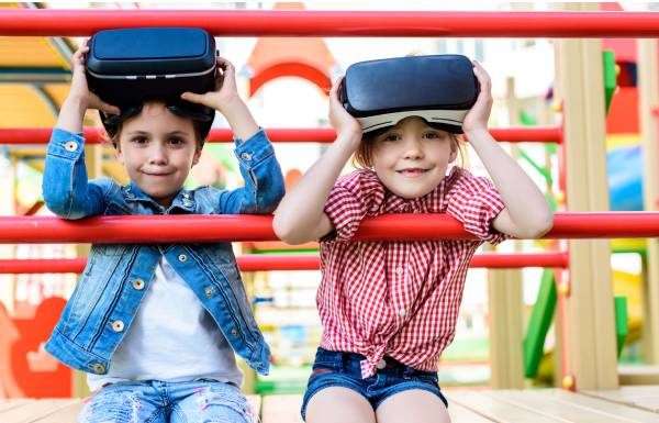 Two kids sitting on playground equipment with VR headsets. The equipment is red and the kids are smiling.