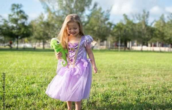 A little girl wearing a purple princess gown. She is standing in a field holding a stuffed animal of a chameleon.