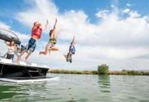 Two boys and a girl wearing life jackets and jumping off the back of a boat into a lake. Their mother is watching from the boat.