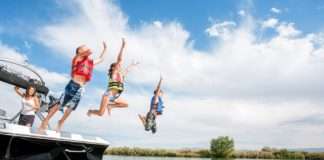Two boys and a girl wearing life jackets and jumping off the back of a boat into a lake. Their mother is watching from the boat.