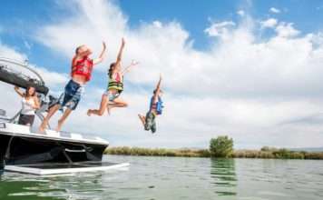 Two boys and a girl wearing life jackets and jumping off the back of a boat into a lake. Their mother is watching from the boat.