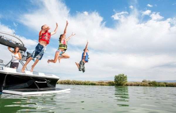 Two boys and a girl wearing life jackets and jumping off the back of a boat into a lake. Their mother is watching from the boat.