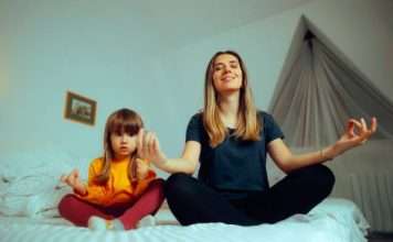 A young mother unwinds by practicing her daily yoga meditations with her daughter on the daughter's bed.