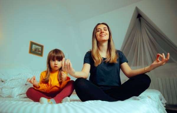 A young mother unwinds by practicing her daily yoga meditations with her daughter on the daughter's bed.