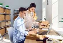 Two people working at a desk with a laptop on it. Several cardboard boxes neatly organized on shelves fill the background.