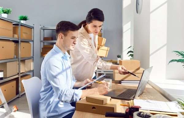 Two people working at a desk with a laptop on it. Several cardboard boxes neatly organized on shelves fill the background.