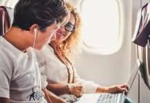 A woman sits by the window of an airplane with a laptop and looks over to a teen boy with headphones.