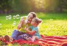 A young woman in a neon green shirt sitting on a red blanket with a little girl in polka dots, blowing bubbles together.