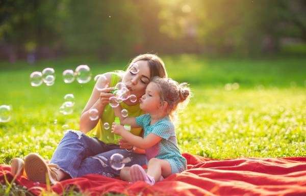 A young woman in a neon green shirt sitting on a red blanket with a little girl in polka dots, blowing bubbles together.