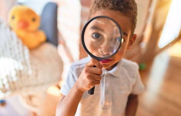 A child holds up and looks through a magnifying glass, which enlarges his eye. A duck toy sits behind him.