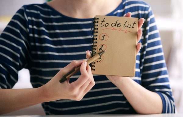 A close-up of a woman holding a notebook page that reads, "To-Do List" and "1, 2, 3." She has a pen pointed to the page.
