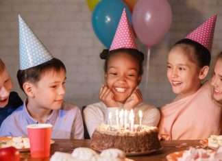 Small children in colorful, polka-dot party hats gathered around a birthday cake with multiple lit candles.