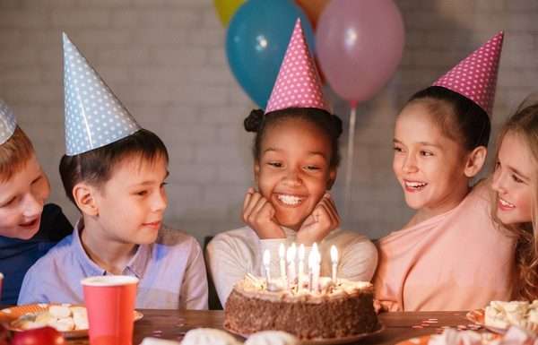 Small children in colorful, polka-dot party hats gathered around a birthday cake with multiple lit candles.