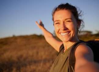 A young, smiling woman with a backpack taking a selfie outdoors and gesturing toward a grassy field.