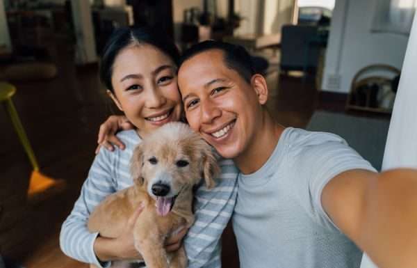 A smiling man and woman holding a happy dog in a warmly lit room with wooden floors and cozy furniture.