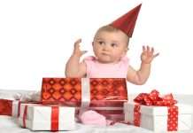 A baby girl wearing a pink set has a red hat on and surrounded by multiple red and white gift boxes.
