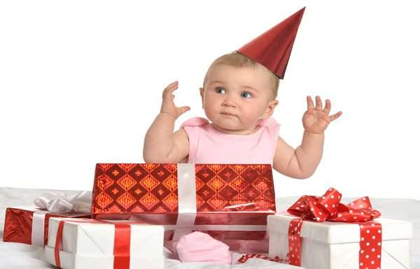 A baby girl wearing a pink set has a red hat on and surrounded by multiple red and white gift boxes.